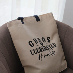 Natúr pamut vászontáska „Chaos Coordinator #momlife” felirattal barna fotelben elhelyezve.