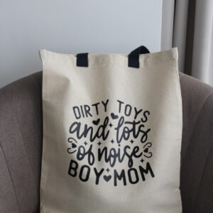 Natúr pamut vászontáska „Dirty toys and lots of noise – Boy Mom” felirattal barna fotelben elhelyezve.