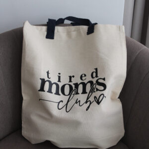 Natúr pamut vászontáska „tired moms club” felirattal barna fotelben elhelyezve.